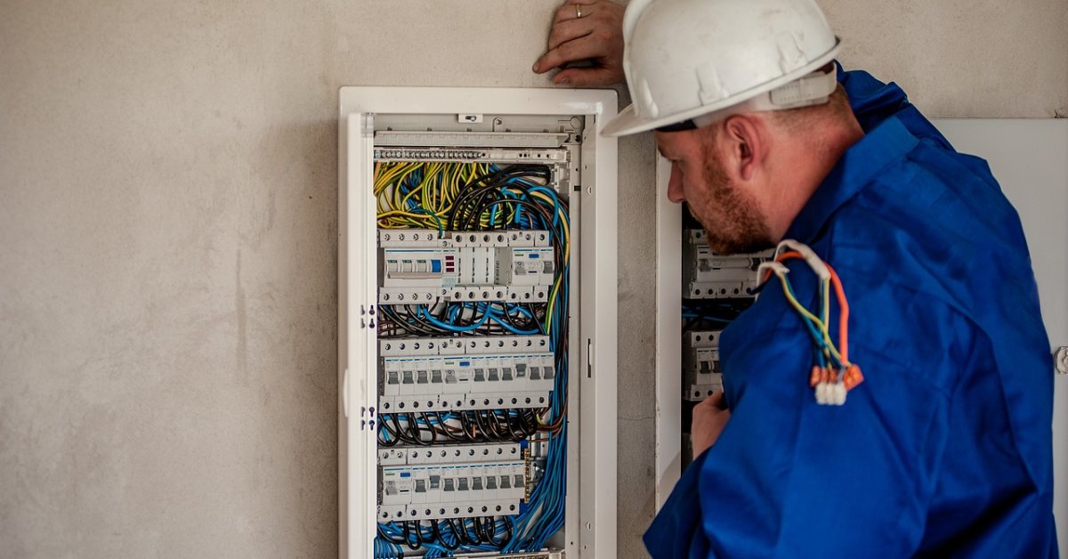 Monteur met helm inspecteert een elektrische verdeelkast met zichtbare bedrading.